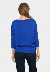 Blue knit sweater with a wide neckline, three-quarter sleeves, and ribbed hem. Worn over dark denim jeans, viewed from the back.