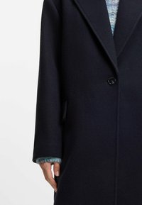 Marinblå ullkappa med noterat krage, enkel knappstängning och en strukturerad blå stickad manschett. Har en sidoficka och rak skärning.