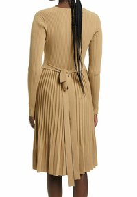 Beige ribbad klänning med långa ärmar, med ett knytbälte i midjan och plisserad kjol. Slät textur, knälängd med rund halsringning.