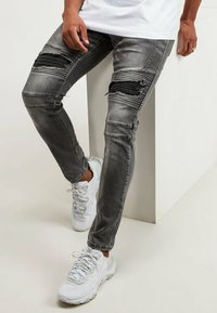 Vaqueros grises desgastados con roturas en las rodillas y detalles en negro, combinados con zapatillas blancas texturizadas. Corte recto con un poco de elasticidad.