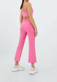 Roze cropped top met kruisende banden, gecombineerd met fitted roze flared broek. Model draagt witte en grijze chunky sneakers. Eenvoudige achtergrond.