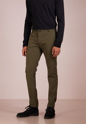 Mand iført olivengrønne slim fit bukser, sort langærmet skjorte og sorte snørebåndssko, der står på en beige gulv.
