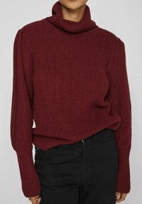 Pull en maille bourgogne à col montant avec texture côtelée, coupe ample et manches longues. Associé à un pantalon noir, affichant un look décontracté.