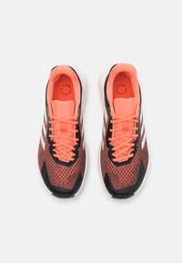 Zapatillas deportivas rojas y negras con una parte superior texturizada, cordones negros y tres bandas blancas. Detalles en naranja neón en la lengüeta y el forro.