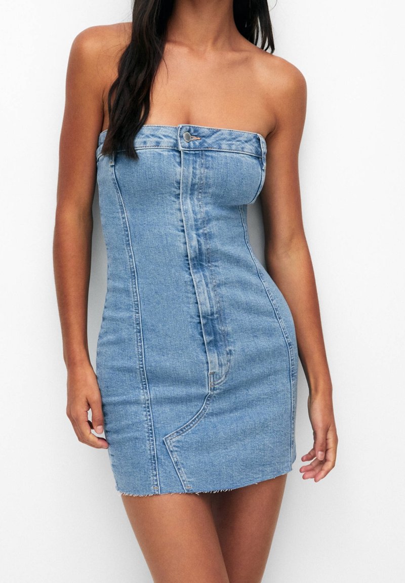 Femme portant une robe mini en jean bleu ajustée sans bretelles avec des boutons sur le devant, debout devant un fond blanc uni.