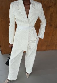 Costume blanc texturé avec un blazer ajusté et un pantalon large, présenté avec une ceinture à la taille et des talons nus pointus.