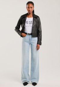 Žena v černé kožené bundě, bílém tričku s potiskem, širokých světle modrých džínách, černém pásku a černých špičatých botách, stojící čelem.