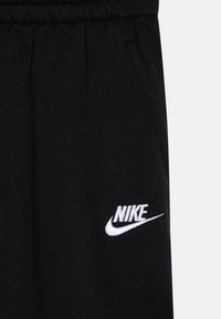 Juodos medvilnės sportinės kelnės su elastingu juosmeniu ir šoniniais kišenėmis. Kairėje pusėje yra balta siuvinėta Nike logotipas.