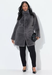 Abrigo de piel sintética gris con un cuello alto, botones visibles y bolsillos laterales, combinado con pantalones negros y un clutch negro. Textura suave.