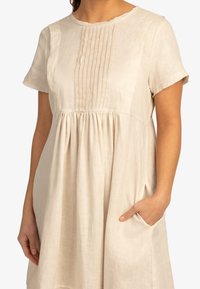 Béžové lněné šaty s krátkými rukávy, skládaným předním dílem, nařaseným pasem a bočními kapsami. Měkká textura s uvolněným střihem.