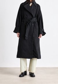 Trench coat in denim nero con bottoni doppiopetto, vita con cintura, maniche larghe e ampio colletto; abbinato a jeans verdi chiaro sfrangiati e stivali neri.