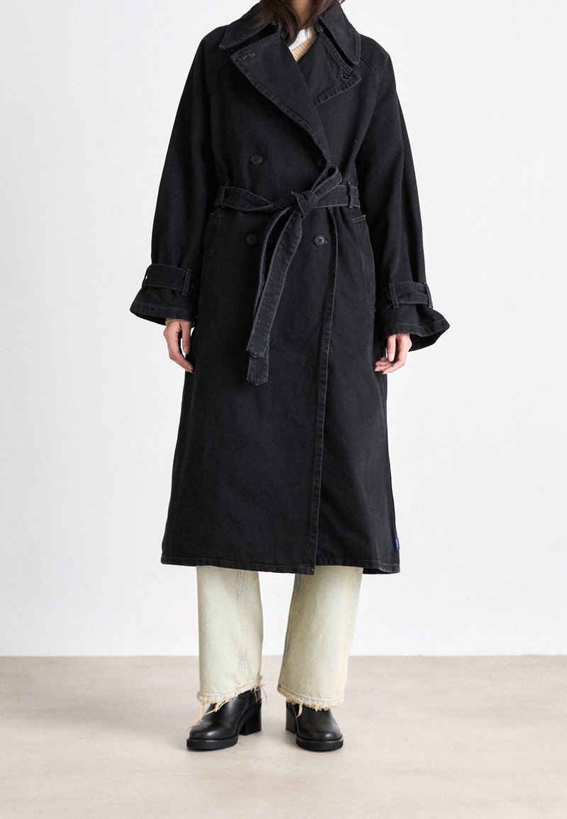 Trench coat in denim nero con bottoni doppiopetto, vita con cintura, maniche larghe e ampio colletto; abbinato a jeans verdi chiaro sfrangiati e stivali neri.