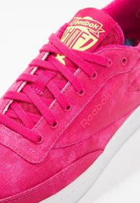 Baskets Reebok rose vif avec lacets assortis, logo jaune sur la languette et semelle blanche, montrées en gros plan sous un angle.