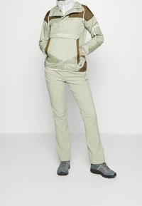 Persona vestida con pantalones verde claro y una chaqueta cortavientos de Columbia a juego con detalles en verde oscuro, combinada con zapatos de senderismo grises.