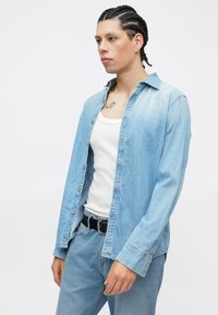 Φωτεινό μπλε πουκάμισο από τζιν με μακριά μανίκια, κουμπωτό σχέδιο, συνδυασμένο με λευκή μπλούζα χωρίς μανίκια και ανοιχτό μπλε τζιν. Εμφανής μαύρη ζώνη.