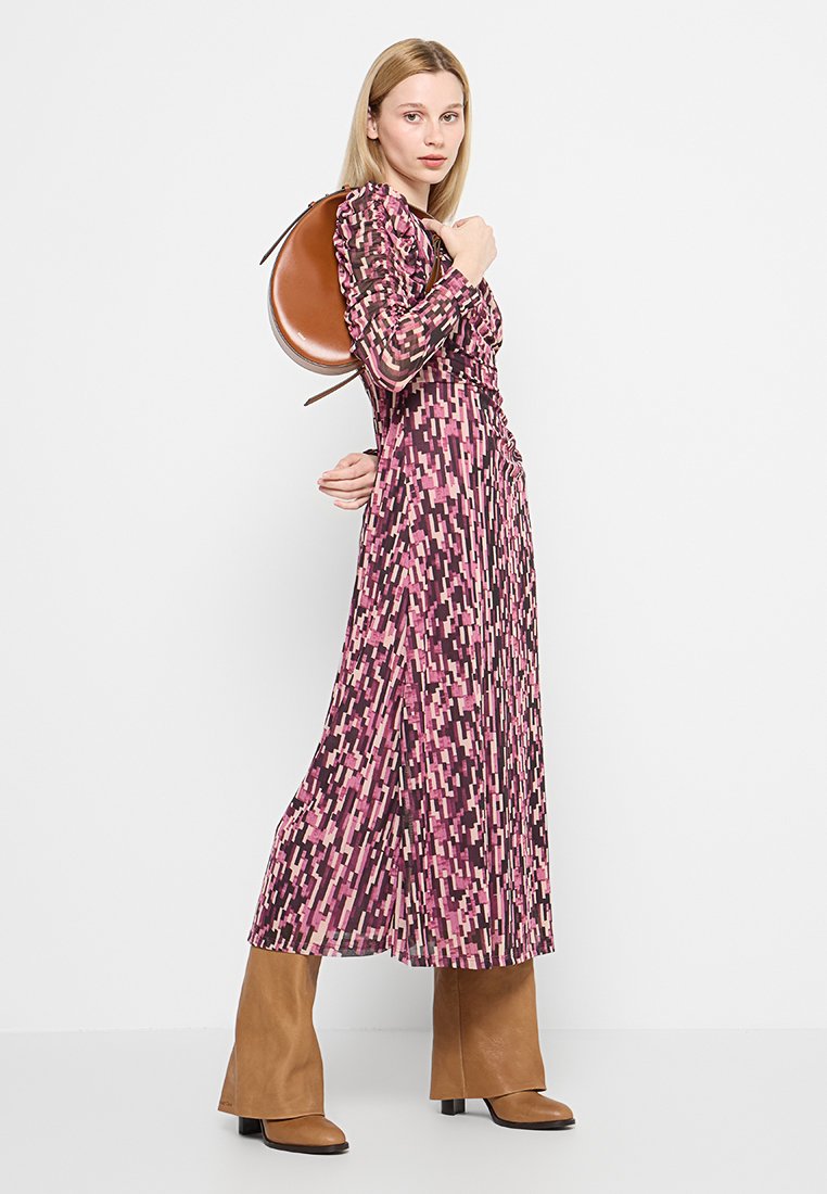 Abito lungo in un motivo geometrico rosa e viola con maniche lunghe, dettagli arricciati e spacchi laterali; abbinato a una borsa circolare marrone e stivali.