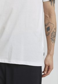 Hvid bomulds t-shirt med korte ærmer, lige kant og et side-tag. En sort tatovering synlig på underarmen tilføjer detalje.