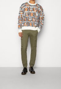Strickpullover mit orange, schwarz und weißen Mustern, kombiniert mit olivfarbenen Cargo-Hosen mit Seitentaschen und schwarzen Schuhen.