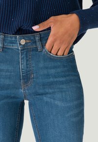 Jean en denim bleu ajusté avec un design à cinq poches, des coutures en cuivre et une fermeture à bouton à l'avant, associé à un pull côtelé bleu marin.