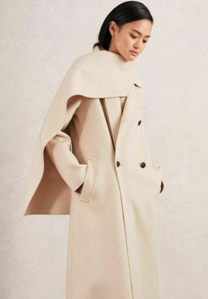 Abrigo de mezcla de lana beige con un cuello tipo capa, cierre de doble botonadura en la parte delantera, bolsillos laterales y textura suave. Diseño minimalista, sin patrones visibles.