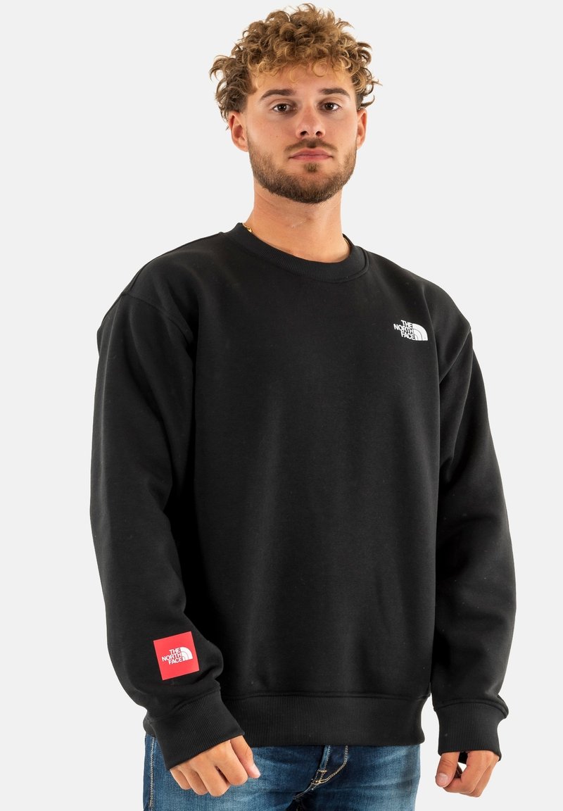 Sudadera negra con cuello redondo, presenta un logo blanco en el pecho y un parche de logo rojo en la manga. Fabricada con un material suave.