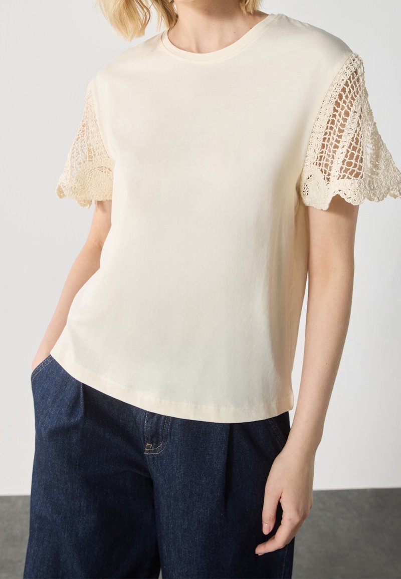 Haut à manches courtes crème avec manches en crochet de dentelle porté avec un pantalon taille haute bleu foncé, main dans la poche, torse visible.
