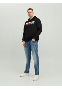 Zwarte hoodie met witte en rode grafische tekst, gecombineerd met lichtblauwe denim jeans en witte sneakers. Model staat voor een neutrale achtergrond.