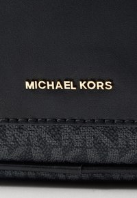 Mustakäsilaukku, jossa on sileä nahka ja teksturoitu kangas, kultainen "MICHAEL KORS" -logo näkyvästi esillä.