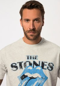 Camiseta de algodón gris claro con un gráfico azul de labios y lengua con el texto "THE STONES" en negro, tipo de impresión vintage.