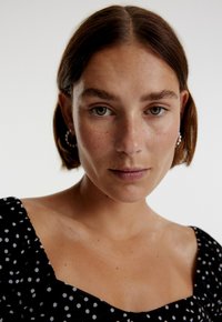 Zwarte jurk met witte stippen en een vierkante halslijn, met pofmouwen. Het model heeft kort haar en draagt ring oorbellen.