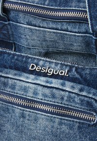 Kék farmerszövet, több cipzáros zsebbel. A "Desigual" márkanevének hangsúlyos nyomata. Textúrált felület, kontrasztos varrással.