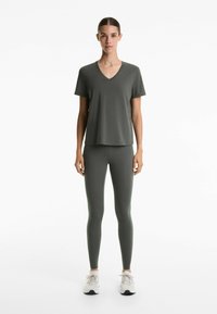 Tricou din bumbac verde închis cu decolteu în V, purtat cu colanți asortați și pantofi sport albaștri. Design simplu, siluetă fitted, textură netedă.