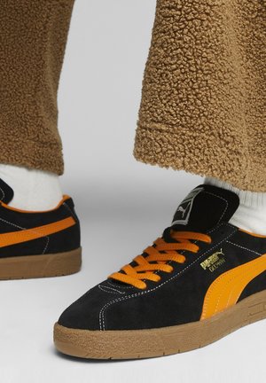 Schwarze und orange Puma-Sneaker mit Gummisohlen, getragen mit weißen Socken und braunen strukturierten Hosen auf einem hellgrauen Hintergrund.
