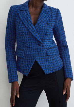 Kvinde iført en figursyet blå og sort hundetandsmønstret blazer med lange ærmer og sorte bukser mod en ensfarvet baggrund.