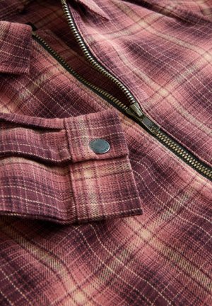 Veste à carreaux vibrante dans des teintes de rose et de bordeaux, en tissu texturé avec une fermeture éclair en métal et des détails de bouton sur le poignet.