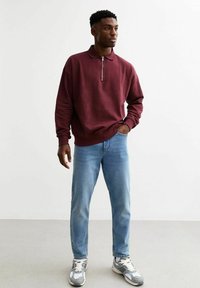 Bordeaux sweatshirt met rits en kraag, gecombineerd met lichtblauwe jeans en grijze sneakers. De stof lijkt zacht en gestructureerd.