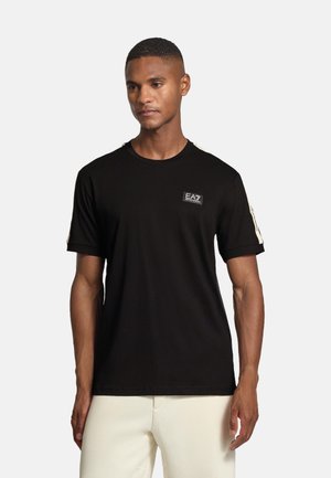 Man som bär en svart EA7 Emporio Armani T-shirt med vit textlogotyp på bröstet och vita randdetaljer på ärmarna, tillsammans med off-white byxor.