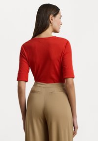 Top rosso aderente con maniche arrotolate, abbinato a pantaloni beige a vita alta con tasche posteriori e una trama di tessuto liscia.