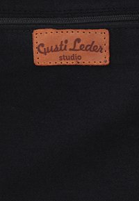 Hnedá kožená etiketa s textom "Gusti Leder studio" prišitá na čiernu látku s viditeľným čiernym zipsom nad etiketou.