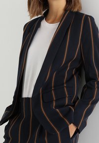 Granatowa marynarka w paski w kolorze brązowym, z zapięciem na jeden guzik i dwoma przednimi kieszeniami. W zestawie z gładką białą koszulką pod spód.