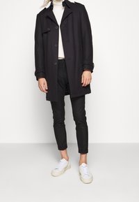 Cappotto di lana nero con una vestibilità su misura, caratterizzato da grandi bottoni, colletto e polsini arrotolati, abbinato a pantaloni neri e sneakers bianche.