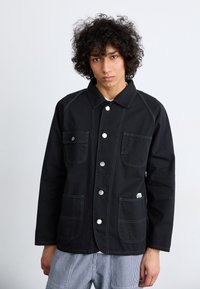 Black cotton jacket featuring two front pockets, silver snap buttons, and contrasting stitching. Simple design with a collared neckline.