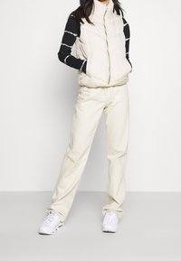 Smanicato imbottito beige con collo alto, chiusura con zip e design trapuntato, abbinato a pantaloni larghi color crema e sneakers bianche. Top a maniche lunghe con righe nere.