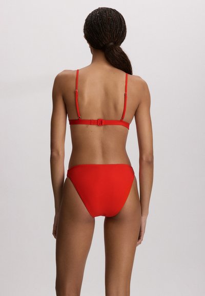 Femeie cu părul împletit într-o coadă joasă, purtând un bikini roșu, văzută din spate pe un fundal deschis simplu.