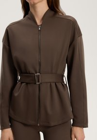 Brun zip-up jacka med hög krage och bälte i midjan, med en slät tygtextur och en strukturerad silhuett.