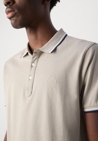 Cămașă polo de culoare bej deschis, cu un material texturat, guler cu două nasturi și dungi contrastante în nuanțe de navy și alb. Prezintă un broderie discretă cu logo.