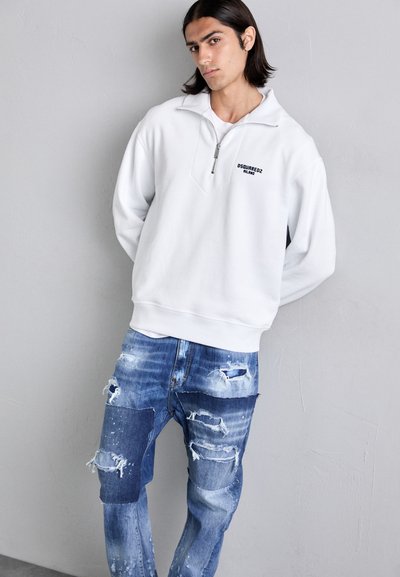 Vit zip-up sweatshirt med en logo på bröstet, tillsammans med slitna blå jeans med lapptäck och fransiga kanter.