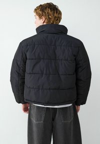 Padded czarna kurtka z wysokim kołnierzem, z widocznymi poziomymi szwami i skróconym fasonem. Połączona z ciemnymi, luźnymi dżinsami.