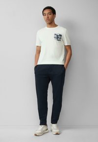 Lichtgekleurde katoenen t-shirt met een marineblauwe grafische zakprint, gecombineerd met donkerblauwe, tapered broek en witte sneakers met bruine accenten.