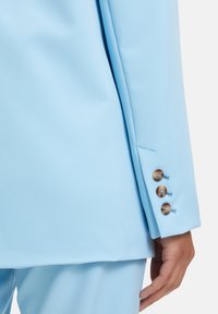 Manche de blazer bleu clair avec trois boutons marron surpiqués de bleu et texture de tissu lisse. Design ajusté avec une coupe structurée.